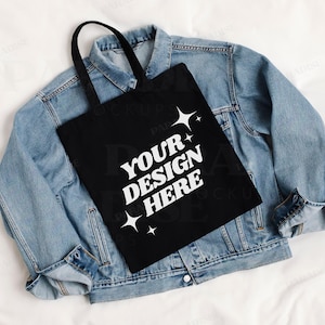 May include: A black tote bag with white text that says "Your Design Here" and stars. The bag is laying on top of a blue denim jacket.