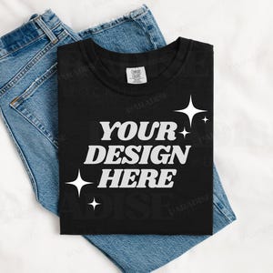 Könnte beinhalten: Ein schwarzes T-Shirt mit dem Text "YOUR DESIGN HERE" in weißen Buchstaben. Das Shirt ist gefaltet und liegt auf einer blauen Jeans.