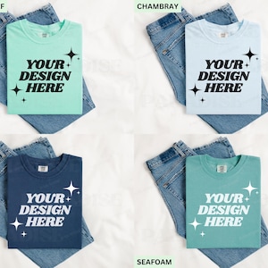 May include: Four t-shirts in various colours: Island Reef, Chambray, Midnight, and Seafoam. Each shirt displays the text "YOUR DESIGN HERE" in white, with starburst graphics. The shirts are folded on top of blue denim jeans.