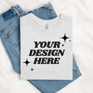 May include: A light gray t-shirt with the text "YOUR DESIGN HERE" in bold black letters, accented by starburst graphics. The shirt is folded and placed on top of a pair of blue denim jeans.