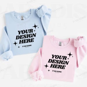 Puede incluir: Dos sudaderas dobladas, una azul claro y otra rosa, con el texto "YOUR DESIGN HERE" en negro. Cada sudadera tiene un gráfico de estrella y la palabra "PARADISE" debajo.