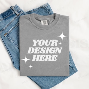 May include: A heather gray t-shirt with the text "YOUR DESIGN HERE" in white, surrounded by small white stars. The t-shirt is laid flat on a white surface with a pair of blue jeans folded underneath.