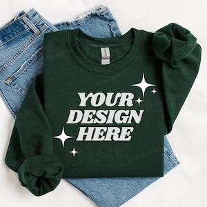 May include: A dark green sweatshirt with the text "YOUR DESIGN HERE" in white lettering and small white stars. The sweatshirt is folded and laid on top of a pair of blue jeans.