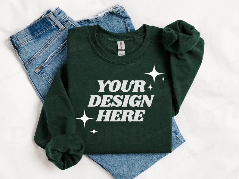 May include: A dark green sweatshirt with the text "YOUR DESIGN HERE" in white lettering and small white stars. The sweatshirt is folded and laid on top of a pair of blue jeans.