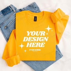 May include: A bright yellow long-sleeved shirt with the text "YOUR DESIGN HERE" in white, along with star graphics. The shirt is folded on top of a pair of blue jeans. The shirt has a black tag.