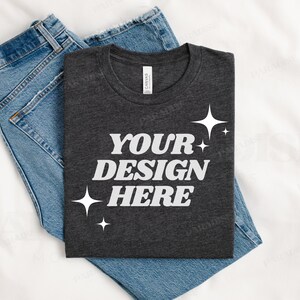 Può includere: Una maglietta grigia scura con il testo "YOUR DESIGN HERE" in lettere bianche e piccole stelle bianche. La maglietta è piegata e posizionata sopra un paio di jeans blu.