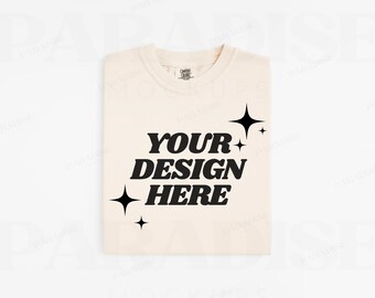 Puede incluir: Una camiseta de color crema con el texto "YOUR DESIGN HERE" en negro. El texto está en un estilo de fuente retro y está rodeado de pequeñas estrellas negras.