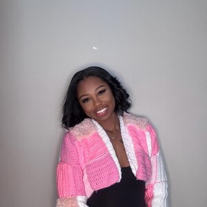 May include: A cropped, patchwork crochet jacket in shades of pink, white, and a touch of faux fur. The jacket features a mix of textures and patterns, including solid blocks, stripes, and a checkered design on the sleeves. The jacket is a unique, handmade garment.