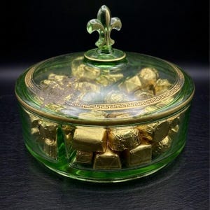 May include: A round, translucent green glass candy dish with a matching lid topped by a fleur-de-lis finial. The lid features a gold-colored Greek key border pattern. The dish is filled with multiple small, square-shaped chocolates wrapped in gold foil.