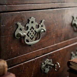 May include: Close-up of a dark brown wooden dresser with three drawers. Each drawer has ornate, antique-style metal handles and keyholes. The wood grain is visible, and the finish is distressed, giving it a vintage look.