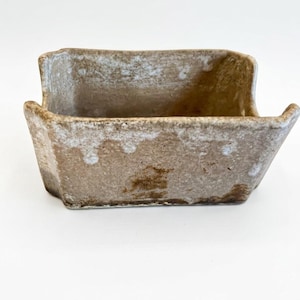 May include: A small, rectangular, ceramic planter with a rustic, weathered appearance. The planter is a muted brown color with white accents along the edges, giving it a textured look. The design features a slightly flared shape with small handles.