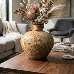 May include: A large, textured, beige vase with a wide, round base and a woven rim, filled with dried flowers. The arrangement includes pink protea, cotton bolls, and various seed pods. The vase sits on a dark wood coffee table.