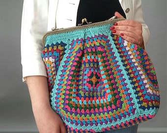 May include: A colorful, handmade crochet bag with a turquoise border and a metal clasp. The bag features a vibrant patchwork design in shades of red, orange, yellow, green, blue, and purple. The bag is held by a person wearing a white jacket and blue jeans.