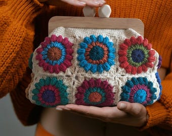 May include: A cream-colored crochet clutch with a wooden frame and white clasp. The clutch features six colorful crochet flower appliqués in shades of blue, red, teal, and purple. The bag is held in front of an orange sweater and pants.