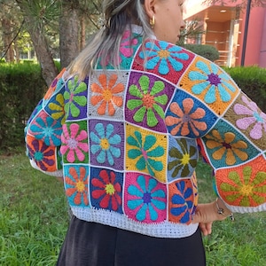 May include: A colourful crochet top with a floral pattern. The top is made of squares with different coloured flowers in each square. The top is white with a white crocheted trim.