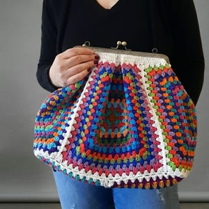 May include: A colorful, handmade crochet bag with a metal clasp. The bag features a vibrant patchwork design in shades of blue, orange, green, and purple, outlined with white crochet. The bag is held by a person wearing a black top and blue jeans.