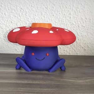 Puede incluir: Una figura caprichosa con forma de hongo, impresa en 3D. La figura tiene un cuerpo azul con pequeñas patas y una cara sonriente. El sombrero del hongo es rojo con manchas blancas y un centro naranja. Sobre una superficie de madera.