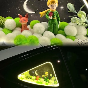 Puede incluir: Luz decorativa para ventana de coche con una escena de "El Principito". La escena incluye una figura del Principito, un zorro y una luna creciente sobre un fondo de noche estrellada. La escena está rodeada de pompones blancos y verdes.