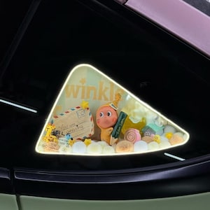 Peut inclure: Un accessoire décoratif pour vitre de voiture avec un personnage de dessin animé, un crayon et une lettre qui dit "You are amazing". L'accessoire est rempli de petits bonbons colorés et d'autres éléments décoratifs. Le mot "wink" est également visible.