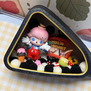 May include: A black, triangular display case with a cartoon figure wearing a pink hat and red overalls. The case contains miniature ice cream cones, Mickey Mouse figures, and a box of Disney-themed treats. The background features a checkered pattern.