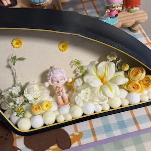 May include: A miniature diorama featuring a small figurine in a pink outfit amidst white flowers, yellow accents, and white pom-poms. The diorama is set in a black frame with a gold trim, creating a whimsical scene.