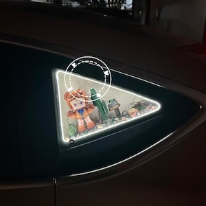 Peut inclure: Une lumière décorative pour vitre de voiture avec un personnage de dessin animé dans une scène de désert, avec des cactus et un panneau indiquant "Lake Mountain". Le personnage porte une tenue orange et un chapeau.