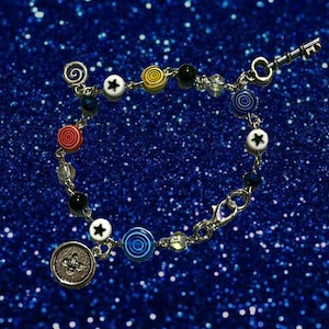 May include: A silver-toned bracelet with a variety of colorful beads and charms. The bracelet features red, yellow, blue, and black circular beads, white star-shaped beads, and clear crystal beads. A key charm and a decorative button charm are also included.