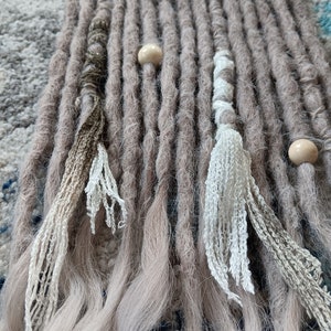 May include: Close-up of several gray and light brown dreadlocks. Some dreadlocks have decorative elements, including wooden beads and white and brown crocheted accents. The dreadlocks are arranged on a textured surface.