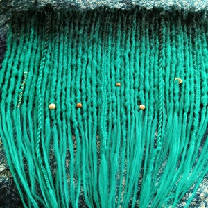 May include: A set of teal and black ombre dreadlocks, adorned with wooden beads and small metal accents. The dreads are arranged in a neat, organized pattern, showcasing a vibrant color gradient. The background is a patterned fabric.
