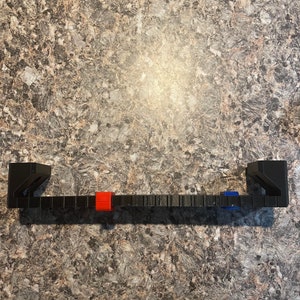 Puede incluir: Un soporte de estante rectangular negro con dos soportes verticales. El soporte tiene una serie de peque&ntilde;as muescas espaciadas uniformemente a lo largo de su longitud. Un cuadrado rojo y azul est&aacute; unido al soporte.