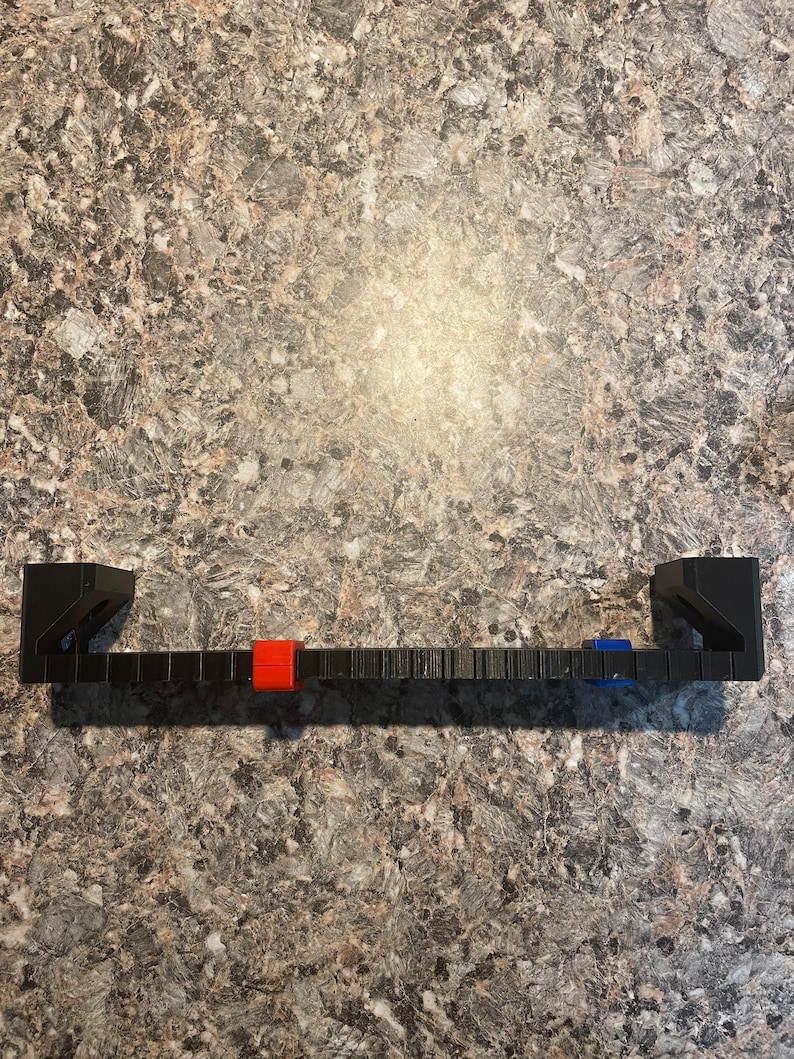 Puede incluir: Un soporte de estante rectangular negro con dos soportes verticales. El soporte tiene una serie de peque&ntilde;as muescas espaciadas uniformemente a lo largo de su longitud. Un cuadrado rojo y azul est&aacute; unido al soporte.