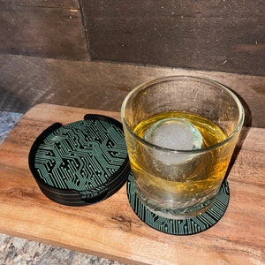 May include: Two black and green circuit board coasters with a glass of amber liquid and a large ice cube on a wooden surface.