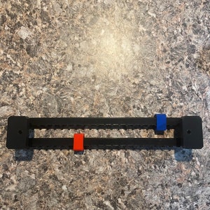 Puede incluir: Un organizador rectangular negro con dos filas de ranuras. Cuenta con una pieza corredera roja y otra azul. El organizador est&aacute; dise&ntilde;ado para sujetar y organizar art&iacute;culos. El fondo es una superficie moteada de tonos neutros.