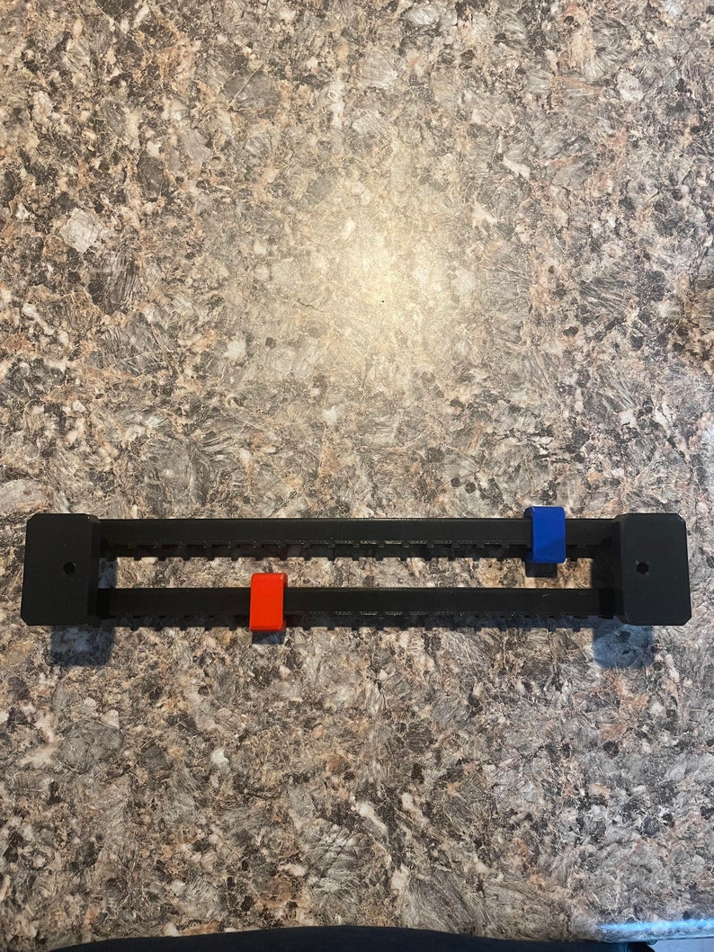 Puede incluir: Un organizador rectangular negro con dos filas de ranuras. Cuenta con una pieza corredera roja y otra azul. El organizador est&aacute; dise&ntilde;ado para sujetar y organizar art&iacute;culos. El fondo es una superficie moteada de tonos neutros.