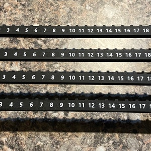 Puede incluir: Dos gu&iacute;as de medici&oacute;n de pl&aacute;stico negro con n&uacute;meros blancos del 0 al 21. Cada gu&iacute;a tiene una serie de ranuras rectangulares y est&aacute; dise&ntilde;ada para medir y alinear elementos. Las gu&iacute;as est&aacute;n sobre una superficie texturizada.