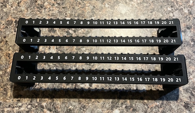 Puede incluir: Dos gu&iacute;as de medici&oacute;n de pl&aacute;stico negro con n&uacute;meros blancos del 0 al 21. Cada gu&iacute;a tiene una serie de ranuras rectangulares y est&aacute; dise&ntilde;ada para medir y alinear elementos. Las gu&iacute;as est&aacute;n sobre una superficie texturizada.