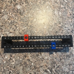 Puede incluir: Una regla negra con n&uacute;meros blancos del 0 al 21. Tiene dos marcadores deslizantes, uno rojo en el n&uacute;mero 7 y otro azul en el n&uacute;mero 16. La regla est&aacute; dise&ntilde;ada para mediciones precisas.