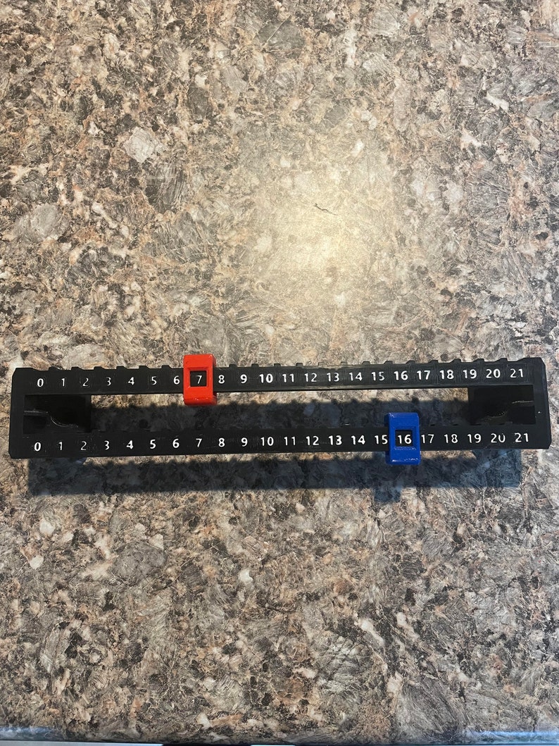 Puede incluir: Una regla negra con n&uacute;meros blancos del 0 al 21. Tiene dos marcadores deslizantes, uno rojo en el n&uacute;mero 7 y otro azul en el n&uacute;mero 16. La regla est&aacute; dise&ntilde;ada para mediciones precisas.