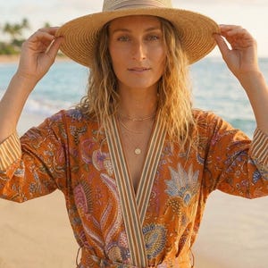 May include: A straw hat with a wide brim and a tan ribbon band, paired with an orange wrap robe featuring a multi-coloured floral paisley print, striped trim along the front opening and cuffs, and a matching tie belt at the waist.