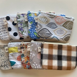 May include: A collection of small, zippered pouches with various fabric patterns. Designs include fish, polka dots, floral, leaves, and plaid. Each pouch has a small tag attached. The pouches are arranged on a white surface.