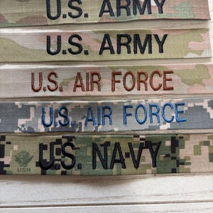 May include: Five rectangular fabric name tapes with embroidered text. Two feature U.S. ARMY in black on camouflage, one U.S. AIR FORCE in brown on camouflage, one U.S. AIR FORCE in blue on digital camo, and one U.S. NAVY in black on digital camo.