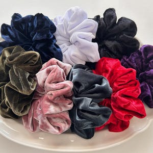 May include: A collection of nine velvet hair scrunchies in various colors including navy blue, white, black, olive green, light pink, charcoal gray, bright red, and deep purple, all arranged together in a cluster on a white scalloped plate.
