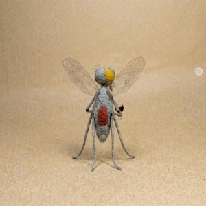 Puede incluir: Una escultura de insecto hecha a mano con cuerpo gris, cabeza amarilla y abdomen rojo. El insecto tiene alas delicadas y transparentes y patas delgadas, sobre un fondo neutro.