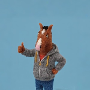May include: A felted horse figure with a brown head, white blaze, and black mane, wearing a gray hoodie, blue jeans, and tan shoes. The figure is giving a thumbs-up gesture. The background is a solid light blue.