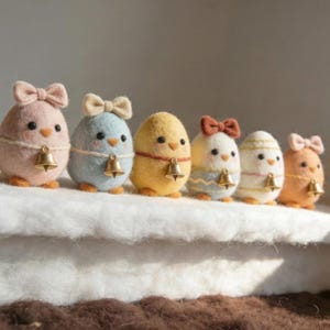 Op de afbeelding: Zes vilten kuikenfiguren, elk in de vorm van een ei met een strik en een gouden bel. De pastelkleuren zijn roze, blauw, geel, wit en oranje. Ze zijn gerangschikt op een wit oppervlak, wat een charmante scène creëert.