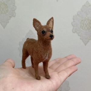 May include: A miniature brown dog figurine with upright ears and a small white spot on its back. The dog is standing on a hand, showcasing its small size. The figurine is made of a textured material, possibly felt or wool.