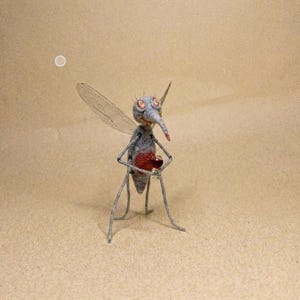 Puede incluir: Una escultura artesanal de un mosquito. El insecto es principalmente gris con un pecho rojo y una larga prob&oacute;scide puntiaguda. Tiene delicadas alas y se apoya sobre finas patas de alambre. El fondo es de color beige liso.