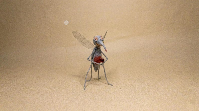 Puede incluir: Una escultura artesanal de un mosquito. El insecto es principalmente gris con un pecho rojo y una larga prob&oacute;scide puntiaguda. Tiene delicadas alas y se apoya sobre finas patas de alambre. El fondo es de color beige liso.