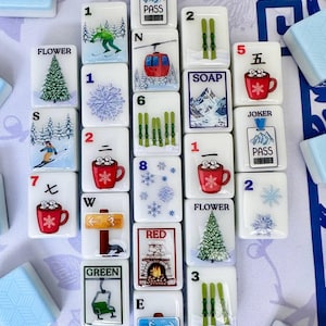 May include: A collection of white Mahjong tiles with winter-themed illustrations. The tiles feature images of skiers, snowflakes, ski lifts, and red mugs. Some tiles have numbers and letters. The text "Joker", "Pass", "Flower", "Soap", "Red", and "Green" are visible.