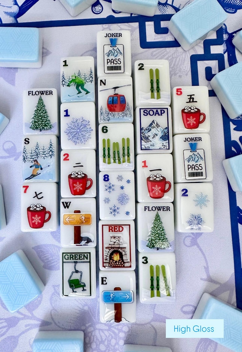 May include: A collection of white Mahjong tiles with winter-themed illustrations. The tiles feature images of skiers, snowflakes, ski lifts, and red mugs. Some tiles have numbers and letters. The text "Joker", "Pass", "Flower", "Soap", "Red", and "Green" are visible.